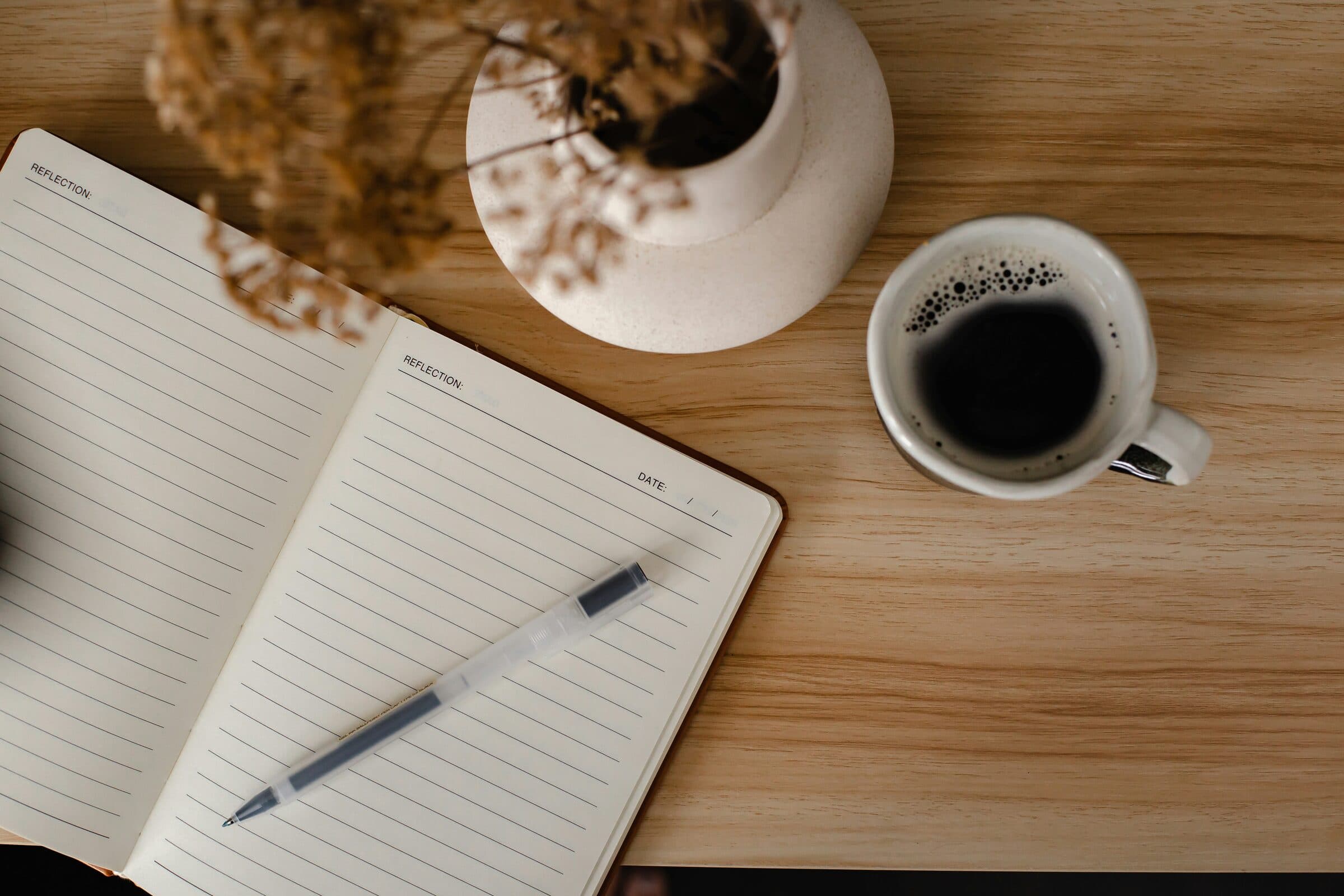 An open lined planner and pen on a wood table, beside a cup of coffee and a vase of dried flowers
