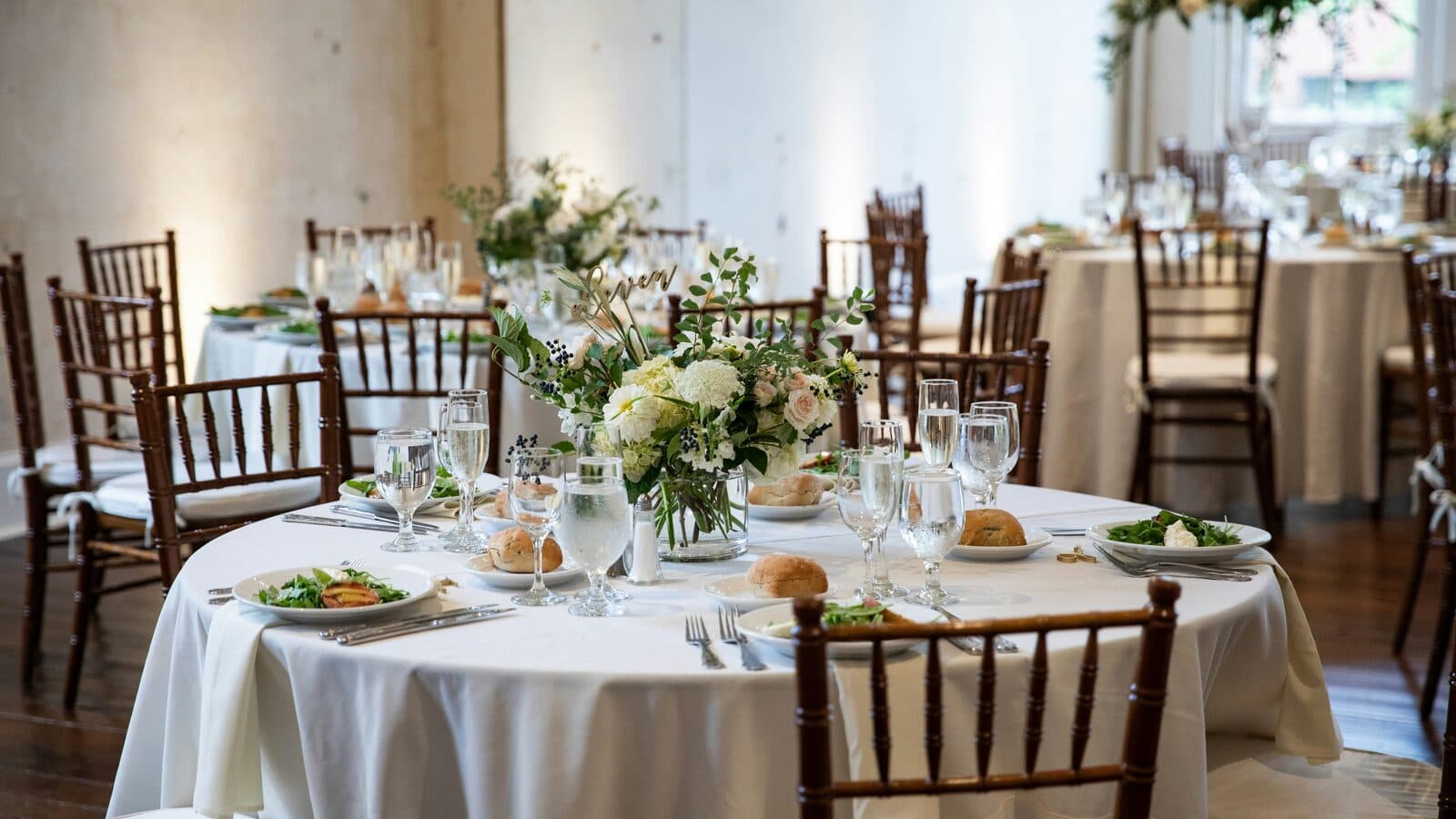 Round reception tables in ivory linen with white and green centerpieces in a bright, plain-walled room