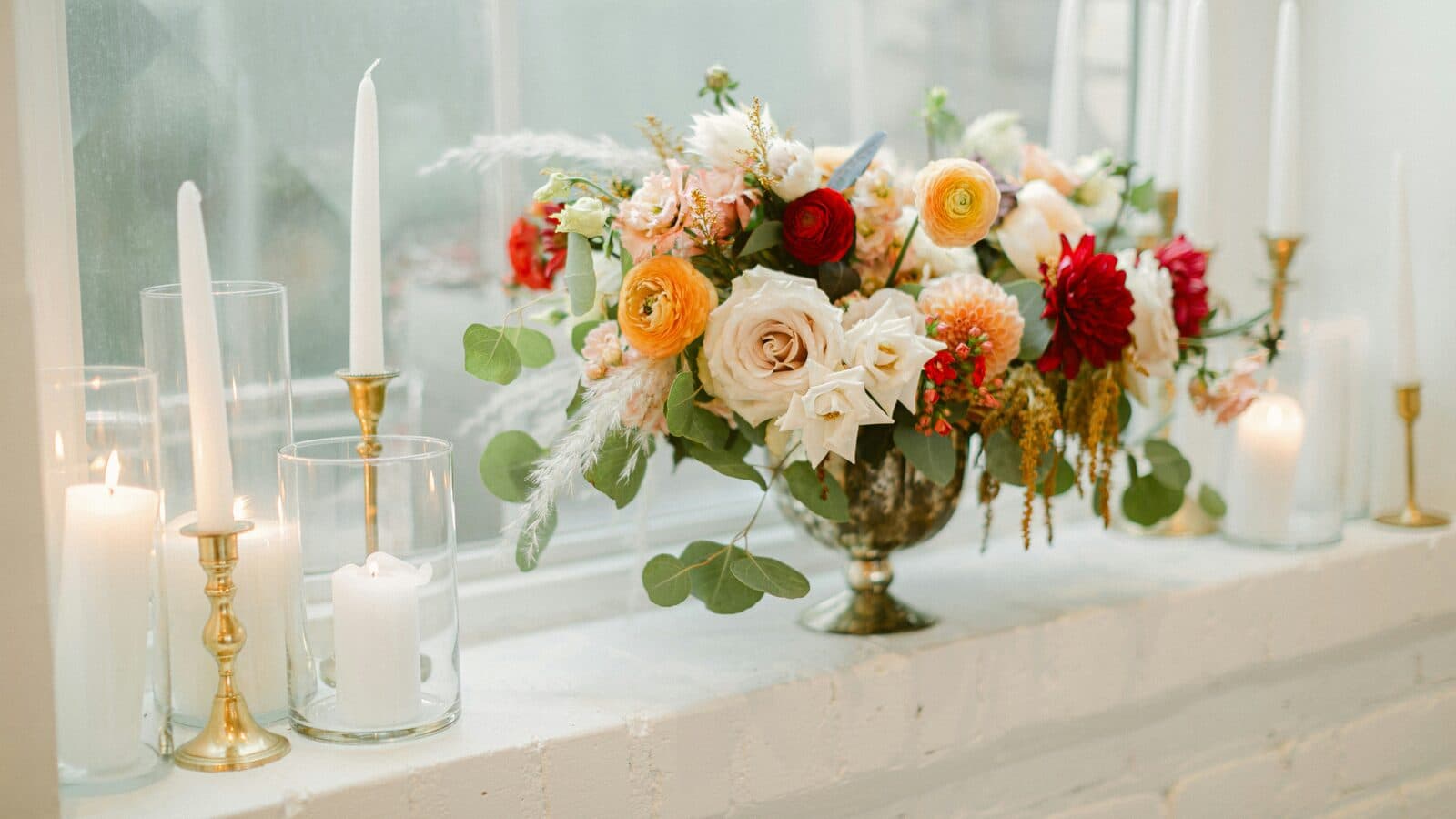 Garden roses in a footed gold urn beside tapered candles in brass holders on a sunlit windowsill