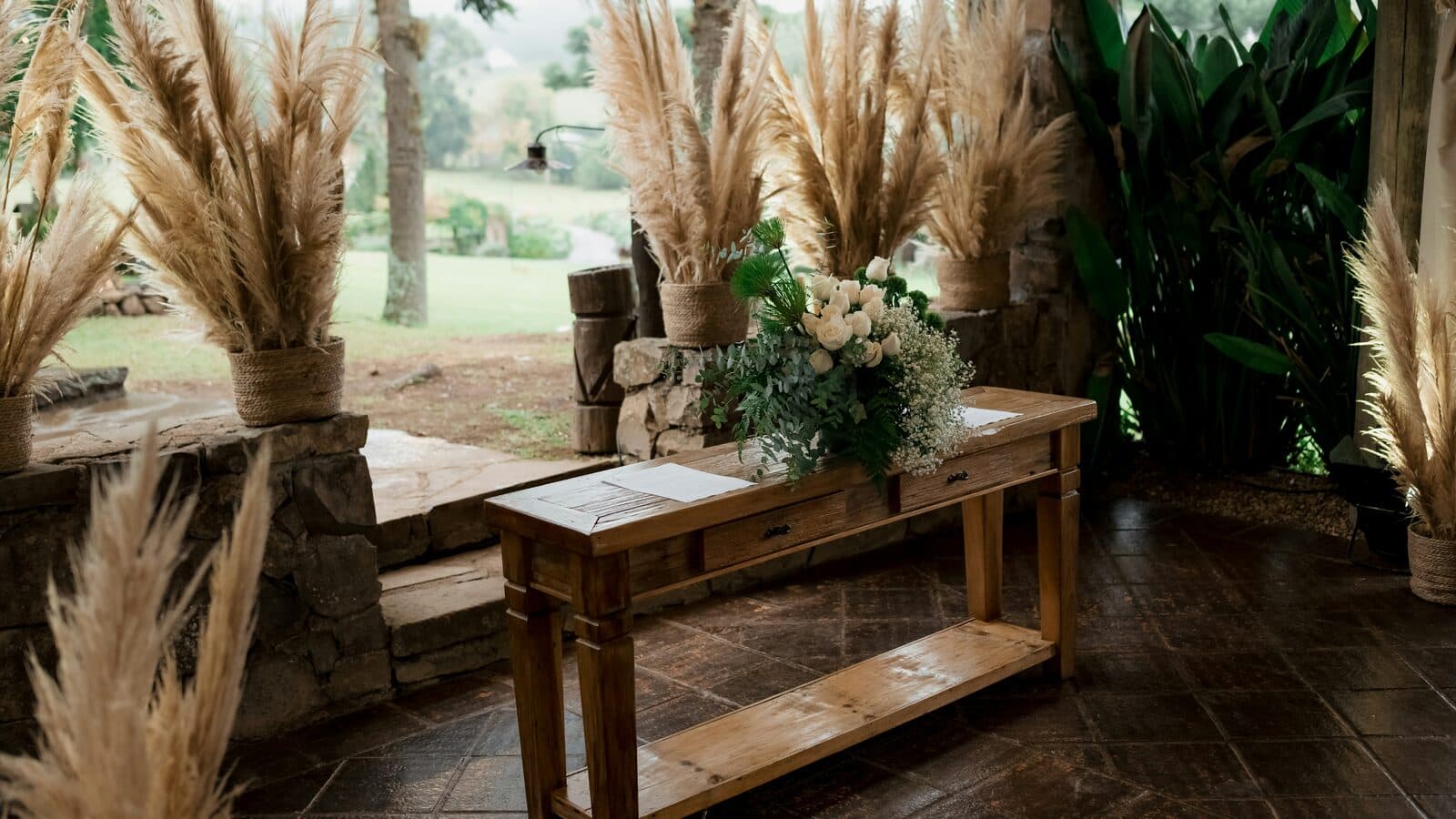 A wooden table with pampas grass and greenery against a stone wall, open countryside behind