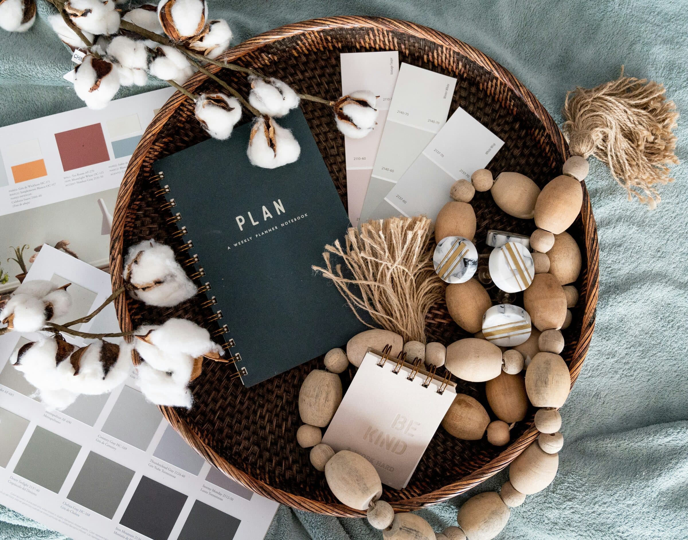 A planning notebook, paint color swatches, cotton stems and wooden beads arranged on a rattan tray